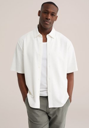 Mand med kort hår iført hvid kortærmet skjorte over en hvid tanktop og grønne bukser, stående med hænderne i lommerne.