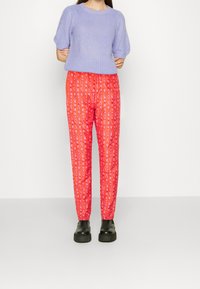 Pull tricot lilas à courtes manches associé à un pantalon rouge vif à motifs avec des rayures verticales et un design graphique. Bottines noires à la cheville.