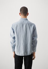 Uomo visto di spalle che indossa una camicia a maniche lunghe blu chiaro e pantaloni neri su uno sfondo chiaro e semplice.