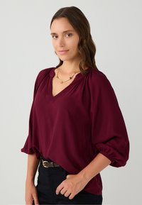 Frau in bluse mit burgunderrot, V-Ausschnitt und Puffärmel, schwarze Hose mit Gürtel und einer kurzen goldenen Kette, die vor einem schlichten Hintergrund posiert.