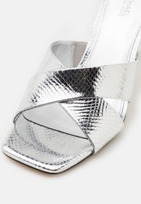 Sandale à talons hauts argentée avec des brides croisées et une texture effet peau de serpent, présentant un bout carré et un design ouvert à l'avant.