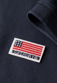 Marineblå sweatshirtstof med et lille broderet mærke med det amerikanske flag og teksten "Superdry" i hvid. Tekstureret materialedetail.