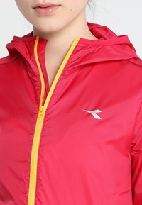 Bright red lightweight jacket with a hood, silver logo accent, and contrasting yellow zipper. Smooth fabric with gathered detailing.
