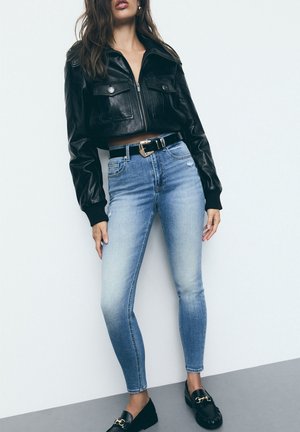 Vrouw draagt een cropped zwarte leren jas, lichtblauwe high-waisted skinny jeans met zwarte riem en zwarte loafers met gouden kettingdetail.