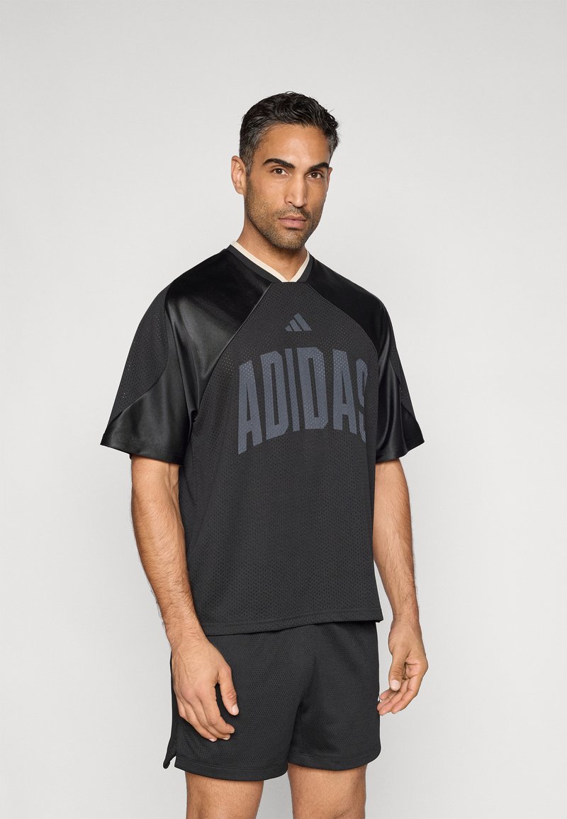 Czarna sportowa koszulka z siateczkową teksturą i krótkimi rękawami, z napisem „ADIDAS” w ciemnoszarym kolorze. W połączeniu z czarnymi szortami, minimalistyczny design.