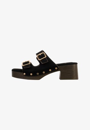Zwarte open-toe sandalen met hak, voorzien van twee verstelbare gouden gespen en gouden studs op een donkeren houten blokhak.