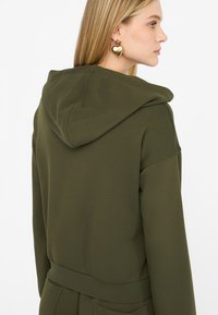 Felpa verde oliva con una texture morbida, con cappuccio regolabile, polsini a coste e vita aderente. Completata da orecchini a forma di cuore dorati.