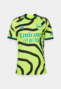 Camiseta de fútbol amarilla neón con patrón de olas negras, mangas cortas, escote redondo, detalles en azul y logo de "Emirates" en el frente.