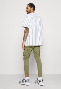Weiß gestreiftes T-Shirt mit kurzen Ärmeln, kombiniert mit olivfarbenen Cargo-Hosen und grauen Sportschuhen. Sichtbare Tattoos und Socken sind ebenfalls zu sehen.