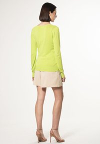 Limettengrüner Pullover mit langen Ärmeln und gerippten Bündchen sowie Saum, kombiniert mit einem beigen Rock. Modell von hinten gezeigt, mit hellen Absatzschuhen.