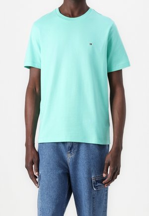 Mann trägt mintgrünes kurzärmeliges T-Shirt mit kleinem Logo auf der Brust und blaue Denim-Cargohosen vor weißem Hintergrund.