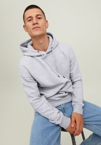 Gray hooded sweatshirt with drawstrings and ribbed cuffs, paired with light blue jeans. Model seated, displaying casual fit and design.