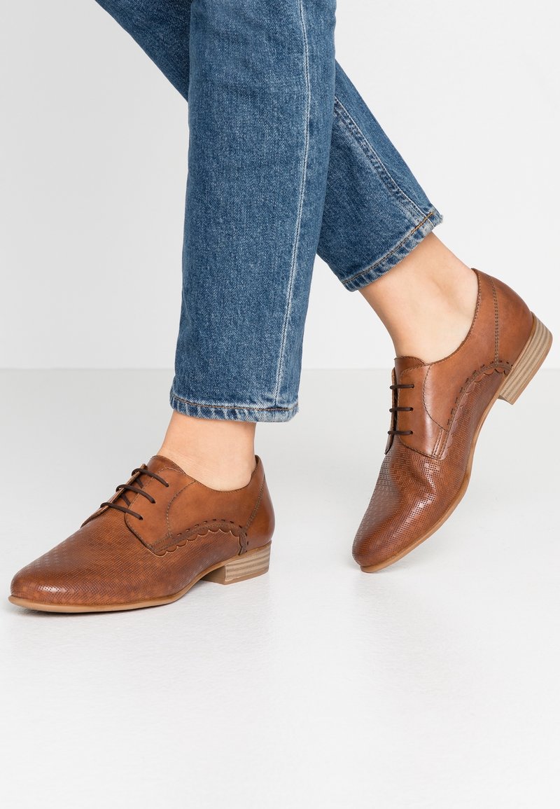 Pieds portant un jean bleu et des chaussures en cuir marron à lacets avec de petits talons en bois, debout sur un sol blanc.