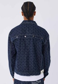 Hombre con el pelo trenzado que lleva una chaqueta de mezclilla oscura y pantalones a juego con patrones de diamantes blancos cosidos, de pie contra un fondo sencillo.