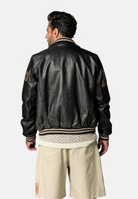 Veste bomber en cuir noir avec col et poignets en tricot rayé. Présente des patches décoratifs sur les manches et un tissu texturé en dessous.