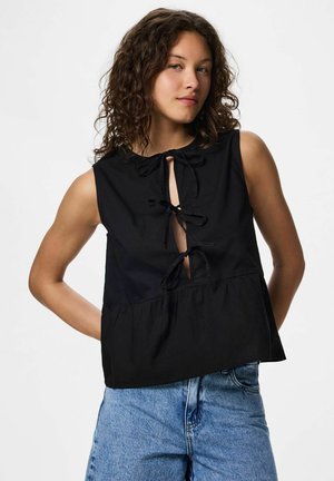 Femme aux cheveux bouclés portant un haut noir sans manches avec trois liens devant et un jean bleu clair, sur fond blanc.