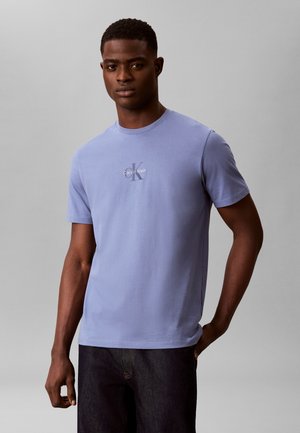 Νέος άνδρας με κοντά σγουρά μαλλιά που φοράει ένα ανοιχτό μωβ t-shirt Calvin Klein και σκούρο παντελόνι, στέκεται μπροστά σε απλό γκρι φόντο.