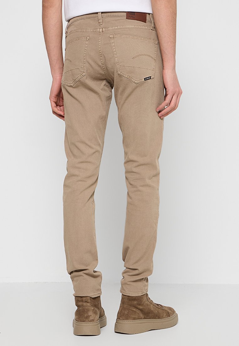 Beżowe spodnie slim-fit o gładkiej fakturze, z dwiema tylnymi kieszeniami i subtelnymi detalami przeszyć. Noszone z brązowymi zamszowymi botkami za kostkę.