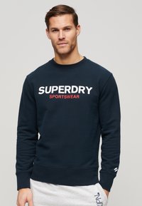Marineblå crewneck sweatshirt lavet af bomuldsblanding, med stort hvidt og rødt tekst "SUPERDRY" og "SPORTSWEAR" på fronten.