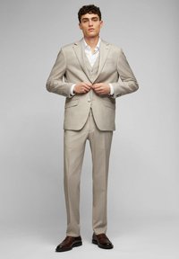 Beige Dreiteiler mit schlanker Passform, unifarbenem Muster, einreihiger Jacke, Reverskragen und Knopfdetails, ergänzt durch braune Schuhe.