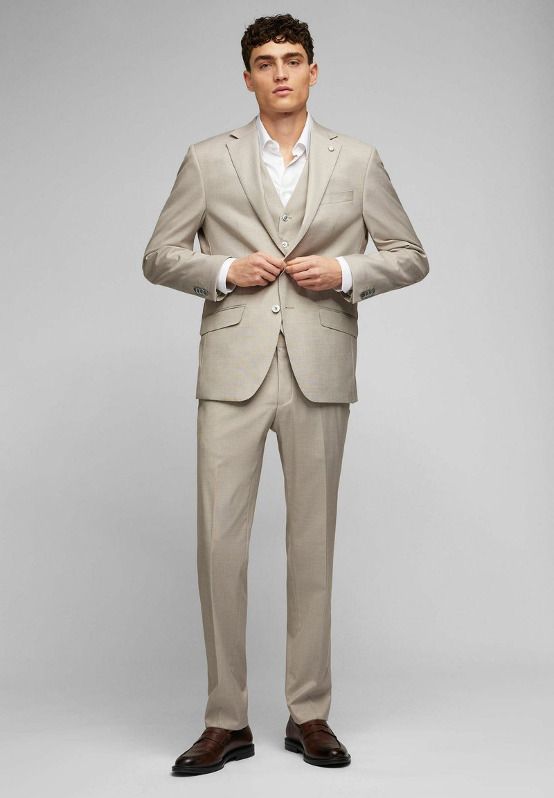 Beige Dreiteiler mit schlanker Passform, unifarbenem Muster, einreihiger Jacke, Reverskragen und Knopfdetails, ergänzt durch braune Schuhe.