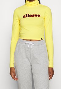 Gelbes langärmliges Rollkragenoberteil mit gesticktem "ellesse"-Logo in Rot, kombiniert mit grauen Jogginghosen mit Kordelzug.