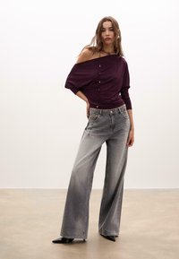 Top bordeaux off-shoulder con dettagli a bottone, abbinato a jeans grigi a gamba larga. Tacchi a punta neri. Sfondo semplice e pulito.