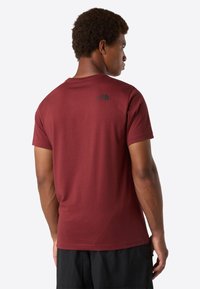 Mann, der mit dem Rücken zur Kamera steht, trägt ein weinrotes T-Shirt von The North Face und schwarze Shorts vor einem hellen, einfarbigen Hintergrund.