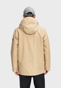 Beige wasserdichte Jacke mit Kapuze, strukturierter Stoff und schwarzem Akzent am linken Ärmel. Das Model trägt eine graue Mütze.