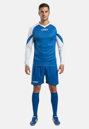 Uomo in piedi che indossa una maglia da calcio a maniche lunghe blu e bianca, pantaloncini blu, calzini blu al ginocchio e scarpe da calcio blu su sfondo bianco.