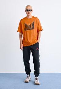 Orange oversized t-shirt med fed sort "JORDAN" tekst, sorte sweatpants med rødt logo, og hvide sneakers med grå detaljer.