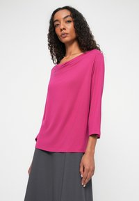 Różowa, luźna bluzka z opadającym dekoltem i rękawami o długości trzech ćwiartek, wykonana z gładkiego materiału, w połączeniu z szarą spódnicą.