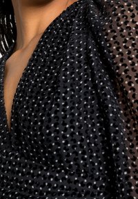 Haut transparent noir avec un motif à pois de différentes tailles, présentant un décolleté en V profond et un détail de tissu texturé. Brillance subtile à la surface.