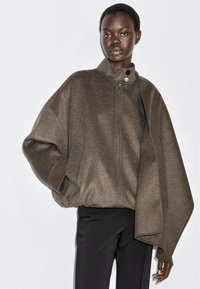 Black woman wearing brown asymmetrical wool jacket with high collar and black pants, posing against plain white background.