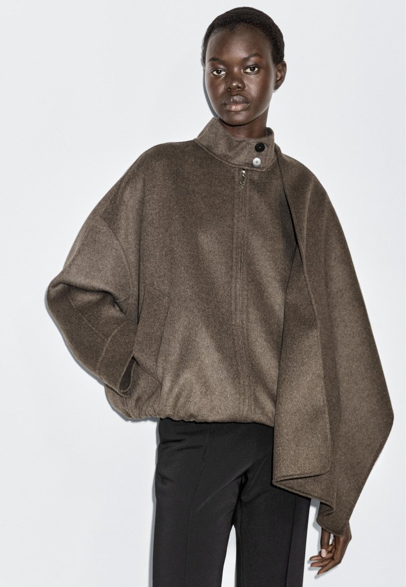 Black woman wearing brown asymmetrical wool jacket with high collar and black pants, posing against plain white background.