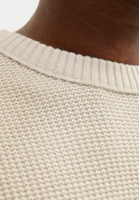 Pull en tricot beige clair montrant un tissu texturé et un col côtelé. L'image se concentre sur la partie arrière du col.