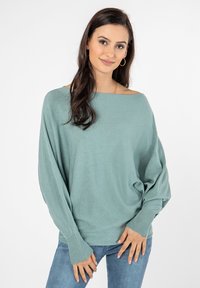 Hellblau oversized Pullover mit weiter Boot-Ausschnitt und gerippten Bündchen. Weiche Textur, getragen über blauen Jeans. Einfaches und lässiges Design.