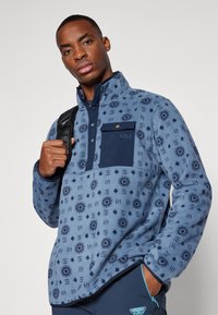Fleece pulover, svetlo modra z mornarsko modrimi akcenti, teksturirana tkanina, vzorčena s sončnimi in diamanti oblikami, vsebuje žep na prsih in visok ovratnik.