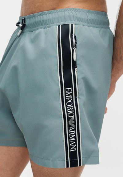 Ανοιχτό μπλε μαγιό σορτς με μαύρη και λευκή κάθετη ρίγα στο πλάι, με το κείμενο "Emporio Armani" και λεπτομέρεια με τσέπη με φερμουάρ.