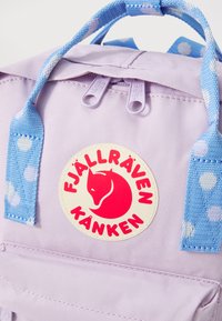 Mochila lilás clara com detalhes em azul com bolinhas, apresentando um logótipo redondo com uma raposa vermelha e o texto "Fjällräven Kånken", além de puxadores de zíper duplos.