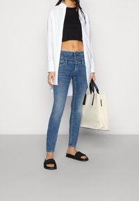 Joven vistiendo jeans ajustados de cintura alta azules, top negro corto, camisa blanca abierta, sandalias negras y sosteniendo un gran bolso de mano beige.