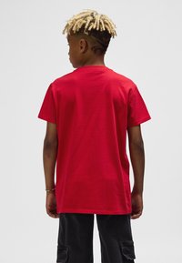 Röd bomulls-t-shirt, korta ärmar, rund hals, avslappnad passform. Sedd bakifrån, bärs med mörka cargobyxor med fickdetaljer.