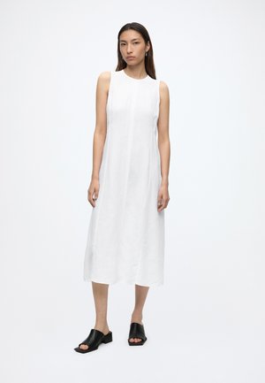 Donna in piedi che indossa un abito midi bianco senza maniche e scarpe nere aperte in punta, su sfondo bianco semplice.