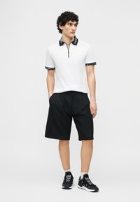 Άνδρας με σγουρά μαλλιά φορά λευκό πόλο με φερμουάρ και μαύρο γιακά, καθώς και μαύρο σορτς, στέκεται με τα χέρια στις τσέπες, κοιτώντας προς τα δεξιά.