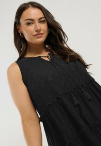 Black sleeveless dress with a textured fabric, featuring a keyhole neck, drawstring ties, and a tiered ruffle hem.