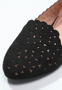 Chaussure plate noire à bout arrondi, présentant un motif floral découpé sur la tige et une doublure intérieure marron clair avec perforations.