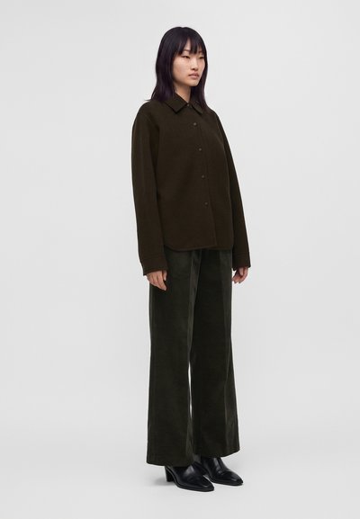 Jeune femme aux cheveux lisses et foncés portant une chemise marron foncé boutonnée, un pantalon large en velours côtelé et des bottines noires à talons, debout de profil.