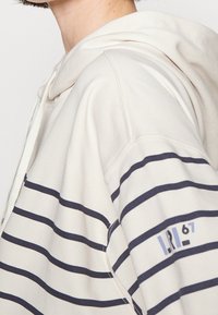 Crème hoodie met marineblauwe strepen, voorzien van een trekkoordkap en een klein logo op de mouw. Zacht materiaal met een relaxte pasvorm.