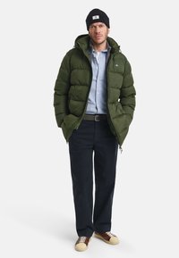 Man in een olijfgroen pufferjack, lichtblauwe button-up shirt, zwarte beanie, donkere broek en bruin-beige sneakers, staande met zijn handen in zijn zakken.