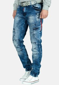 Jeans in denim blu con vestibilità slim, dotati di numerose tasche, dettagli cuciti e una cerniera rossa. Abbinati a sneakers bianche.